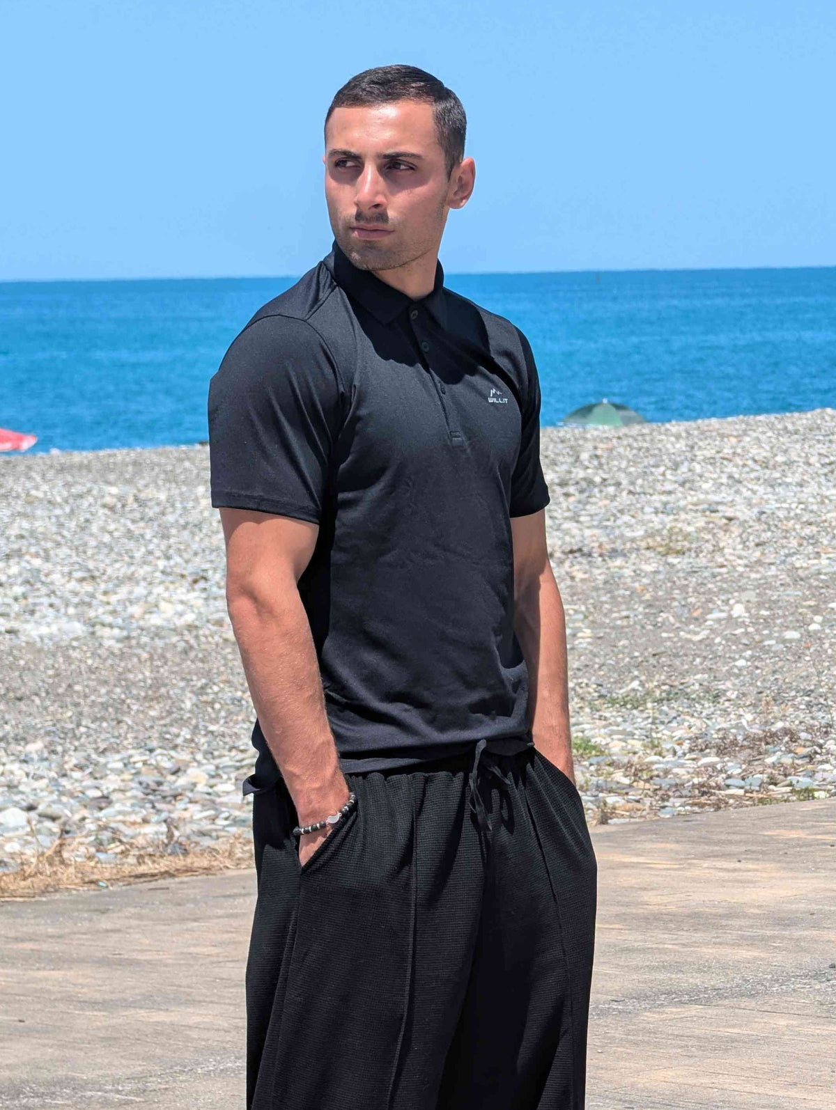 Man in black outfit standing on a wooden boardwalk with ocean and clear blue sky in the background