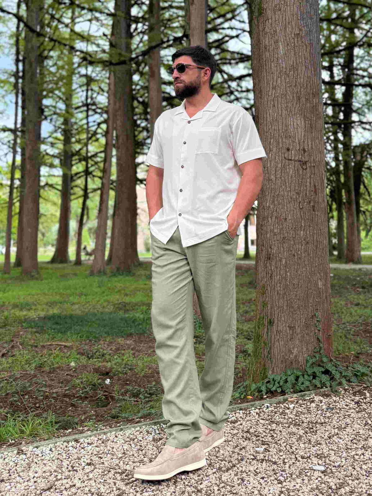 Man standing in a forested area wearing sunglasses and casual clothing.