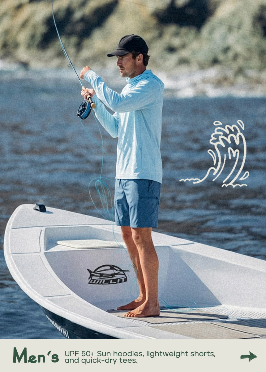 A man stands barefoot on the deck of a boat fishing, wearing WILLIT light blue long-sleeve sun hoodie, blue aathletic shorts, and a black cap.