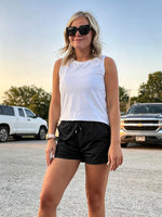 A woman wearing a white sleeveless crop top and WILLIT 2.5 inch black athletic shorts stands in a parking lot at sunset 