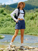 A woman stands on rocks by a stream in a lush green outdoor setting, wearing a beige wide-brim hat, a white long-sleeve shirt tied at the waist, and navy blue hiking shorts 