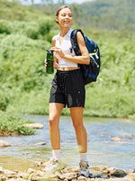 A woman hiking outdoors in a grassy, wooded setting, wearing lightweight cargo shorts 