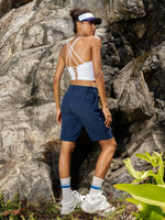 A woman stands outdoors against a rocky cliff wearing a white strappy sports bra, navy athletic shorts_Women's Outdoor Cargo Shorts 10 Inch 